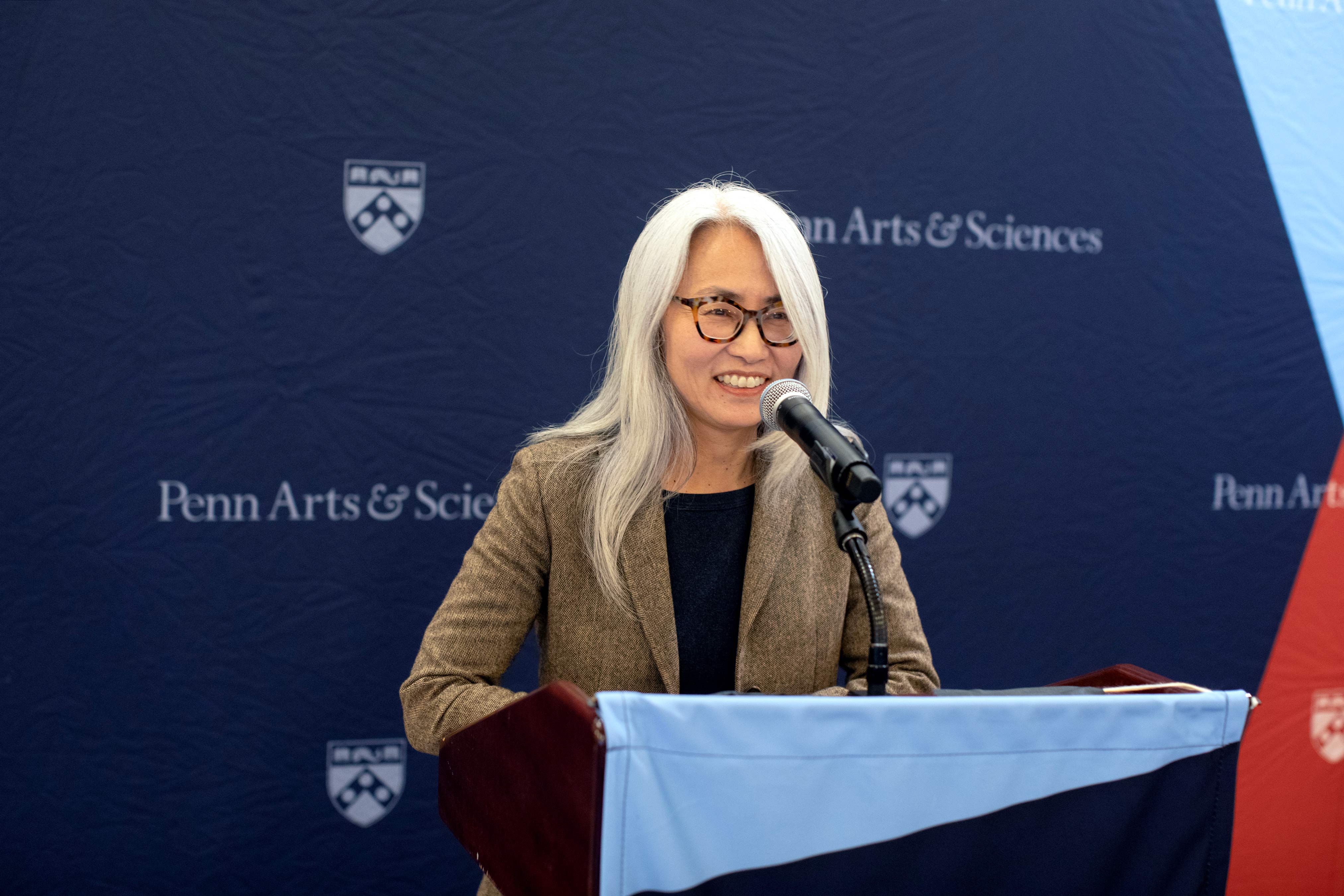 Josephine Park standing in front of a podium and speaking into a microphone, smiling. Behind her is a backdrop with the Penn shield and "Penn Arts & Sciences" repeated in a pattern.