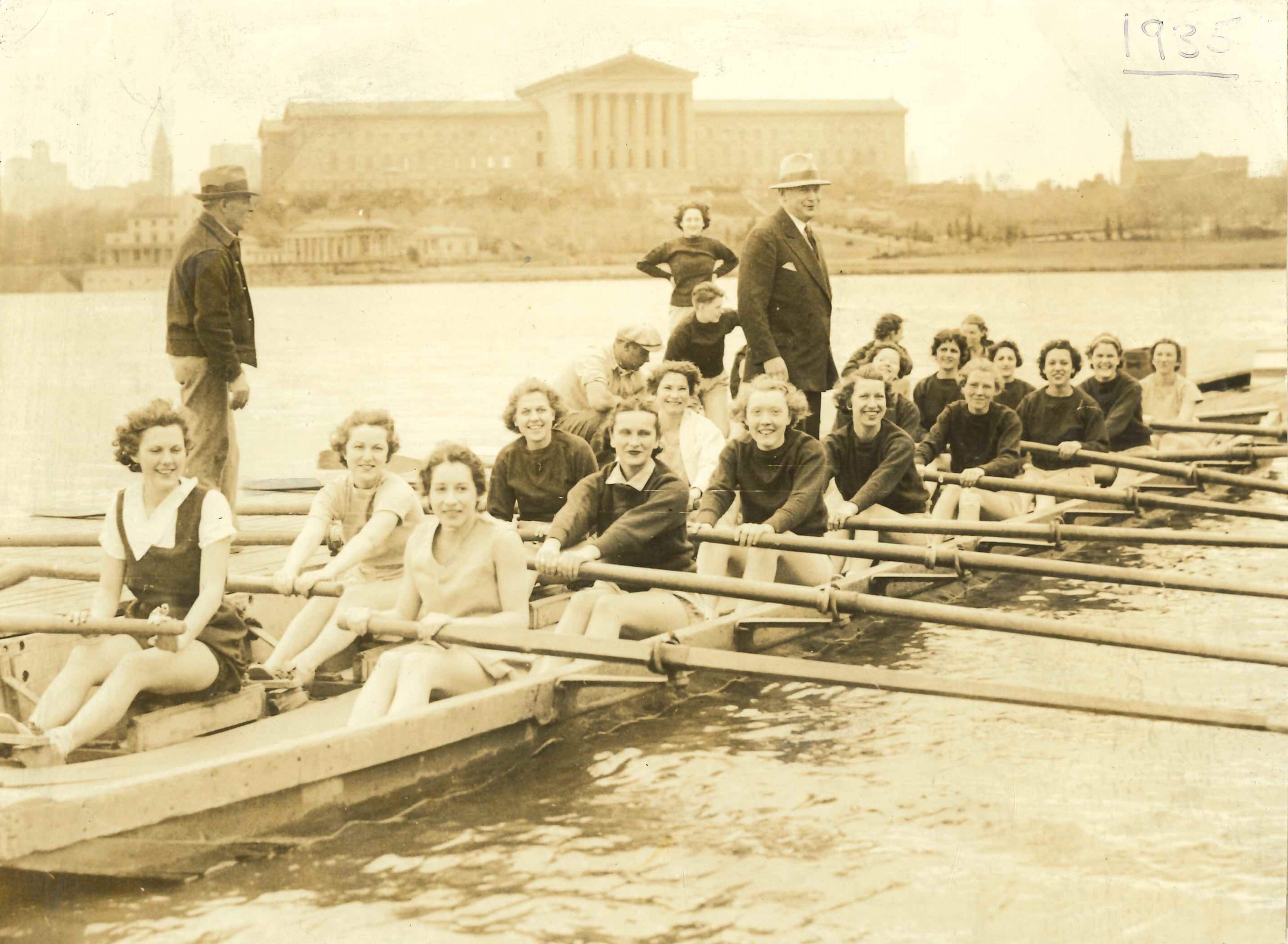 A crew team made up of women on the Schuykill River moored at a dock, with several people standing or crouched behind them. In the background is the Philadephia Museum of Art. in the top right is the underlined number "1935", handwritten and faded.