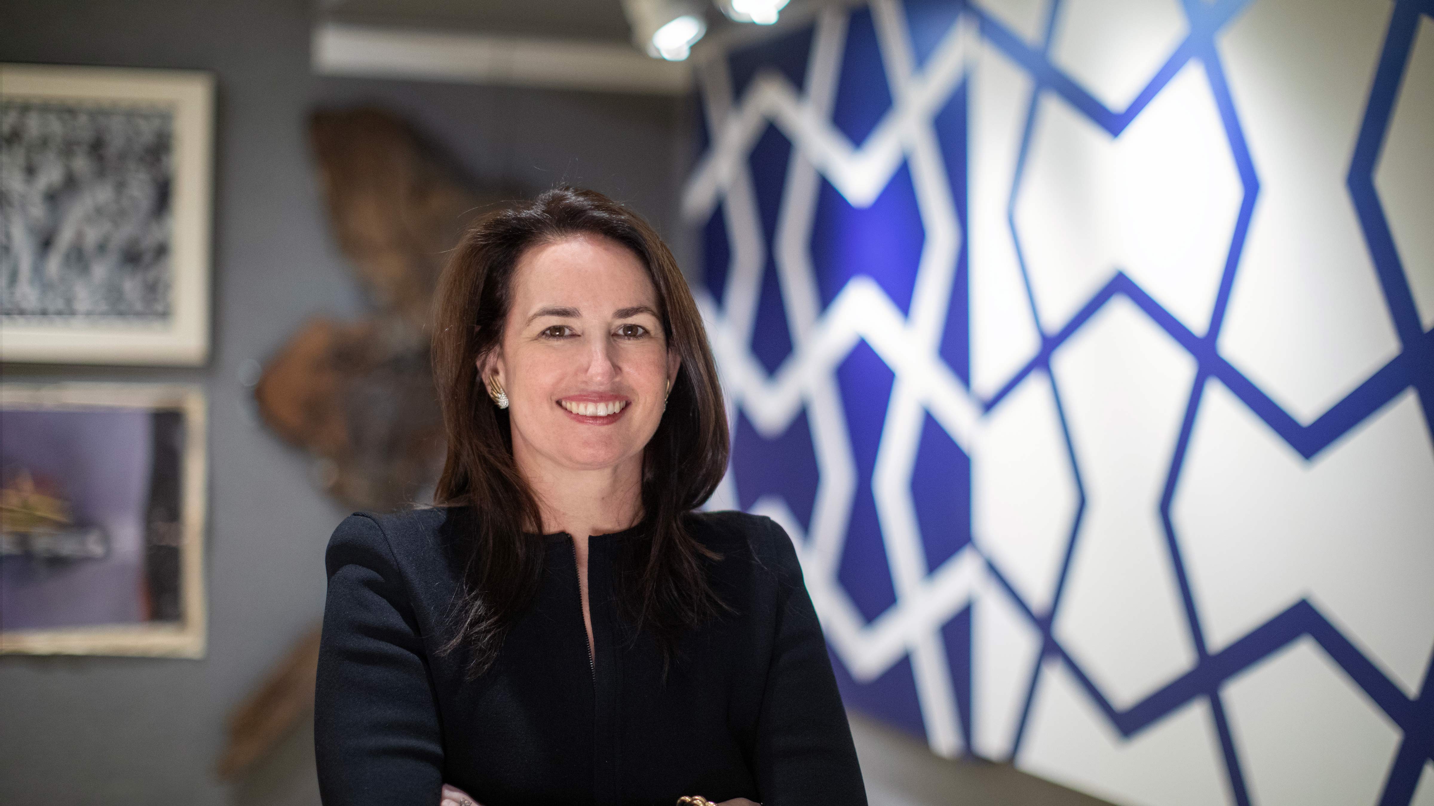 Laura Doyle, standing in an art gallery. Behind her is a geometric patterned wall, and she is smiling.