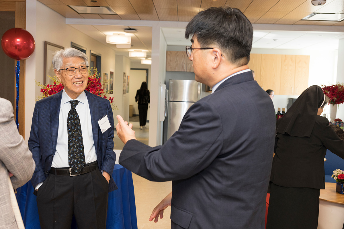 One person facing the camera, hands in pockets, smiling, listening to another person whose back is to the camera. They are engaged in what seems like lively conversation.