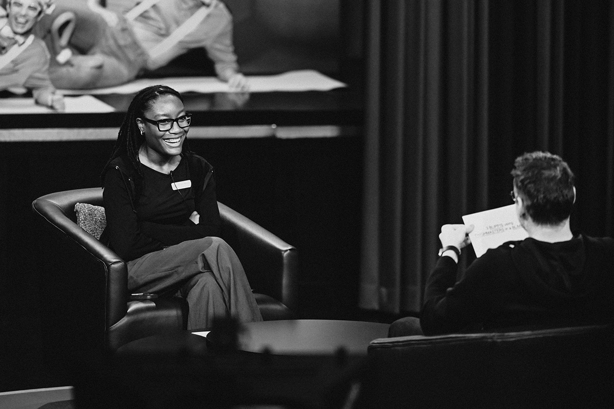 Crystal Marshall on the set of The Tonight Show sitting in a chair laughing. A person is sitting across from her holding a piece of paper. Behind her are people or statues laying on a stage.