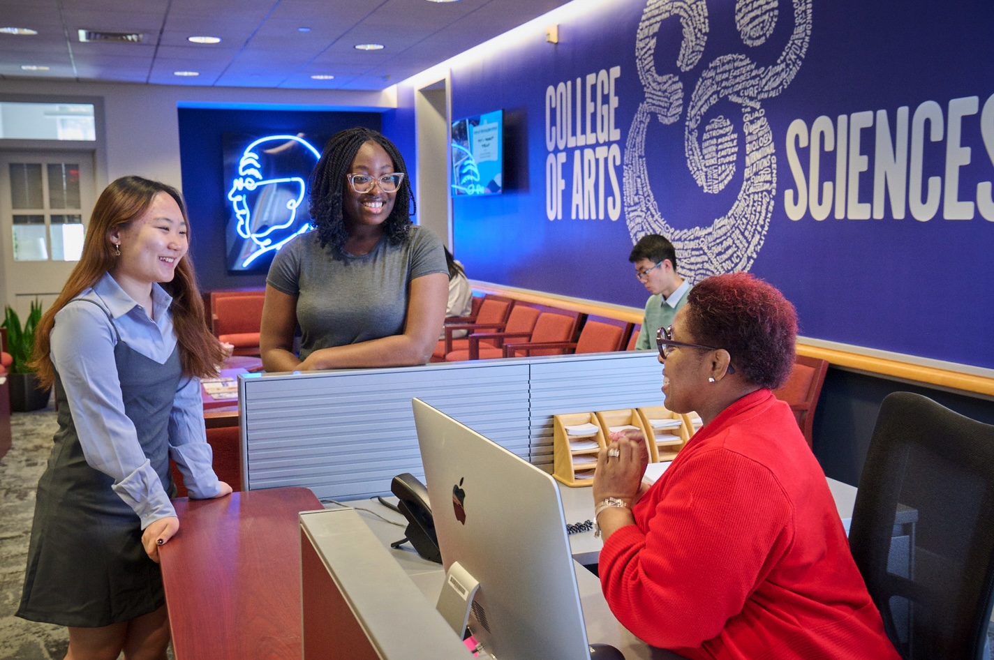 Two students standing in front of a desk where a woman sits in front of a computer. 