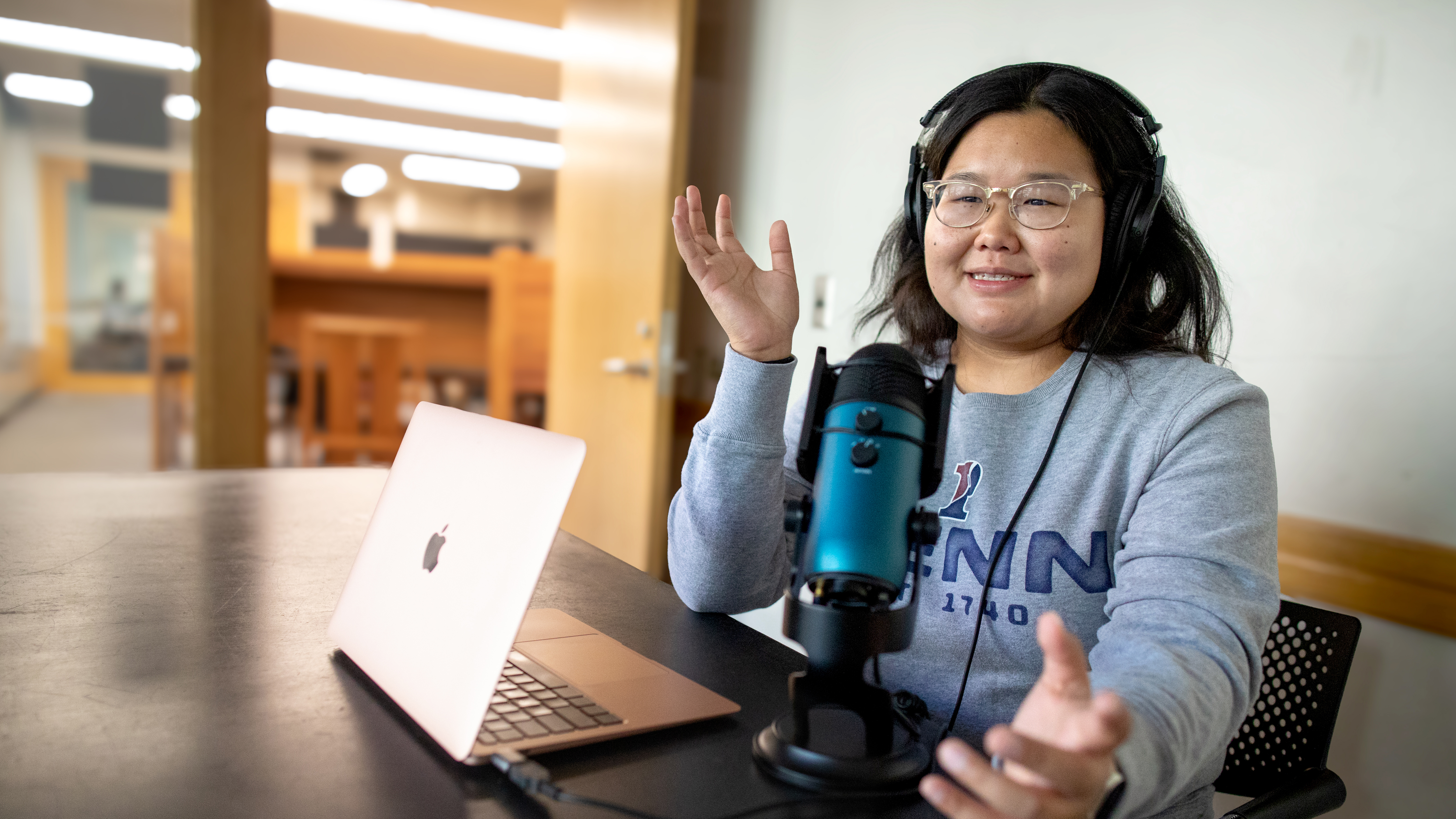 A person sitting at a table wearing headphones, a microphone and computer open in front of her. She is gesturing with her hands as if she is speaking.