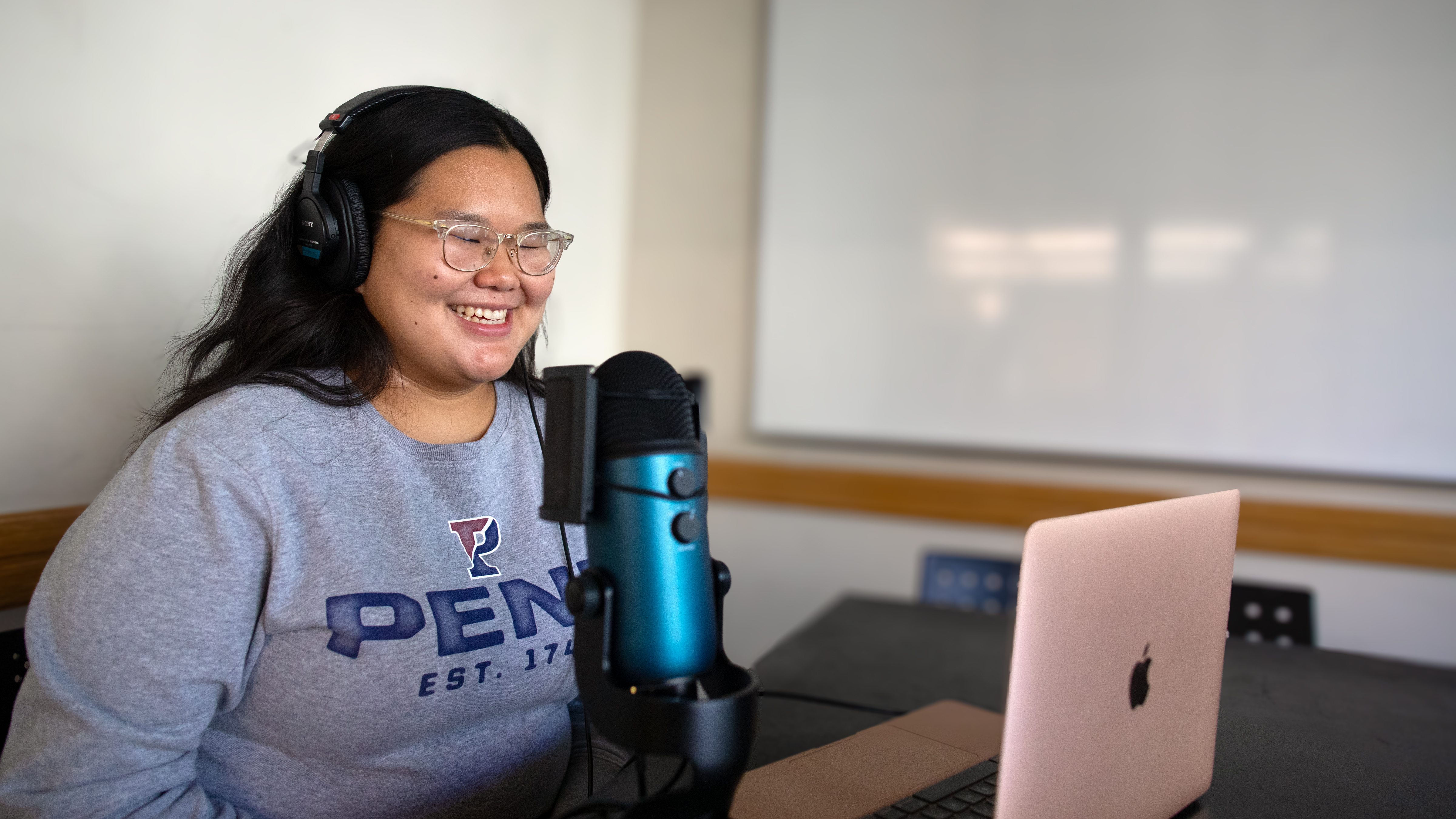 A person sitting at a table smiling, wearing headphones. A microphone and open computer sit at the table in front of her.