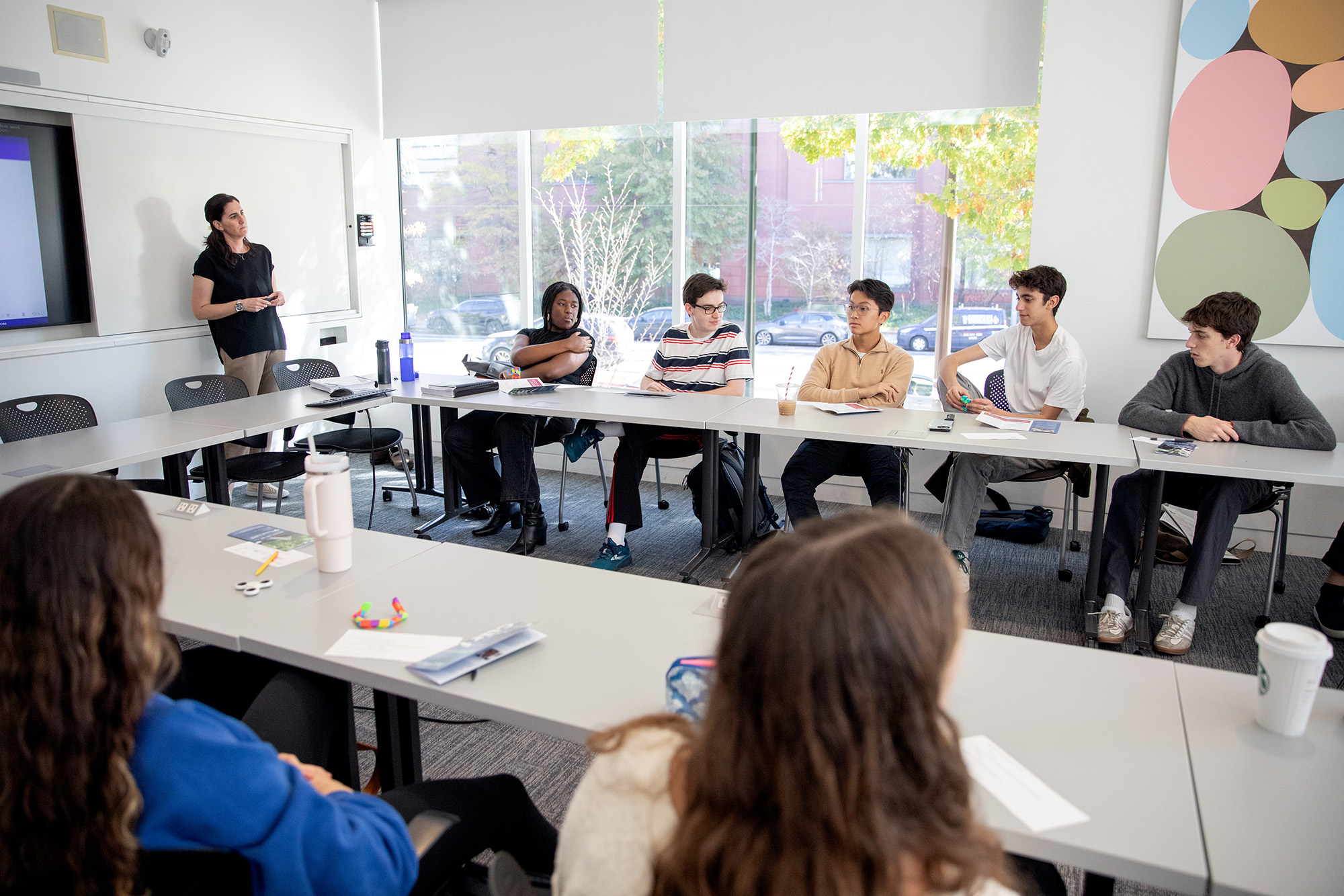 A class sitting around a U-shaped table, with windows to the outside behind the students on the far-side of the table. Michele Margolis, the instructor, is on the far-left of the image. 