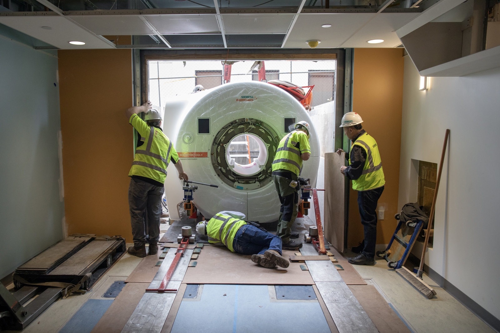 An fMRI machine being installed inside of a building, with four people in yellow construction vests working on the install.