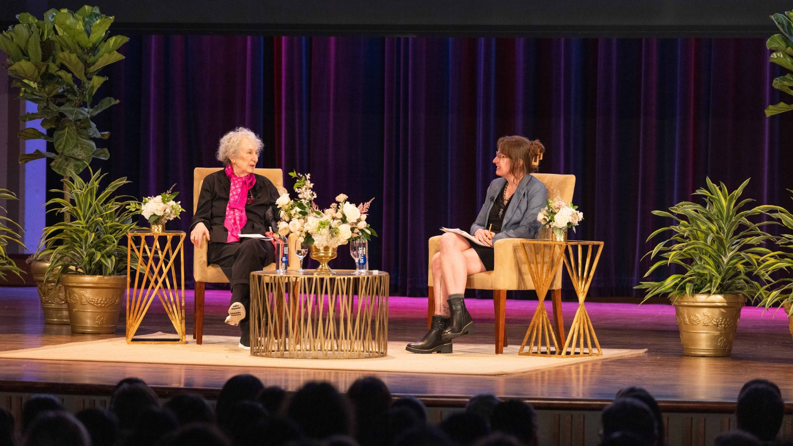 Margaret Atwood and Emily Wilson sitting on a stage in front of an audience. They're making eye contact and speaking to one another with pleasant expressions. Around them are various potted plants, and in front of them is a table with a boquet of flowers, water bottles, and drinking glasses.