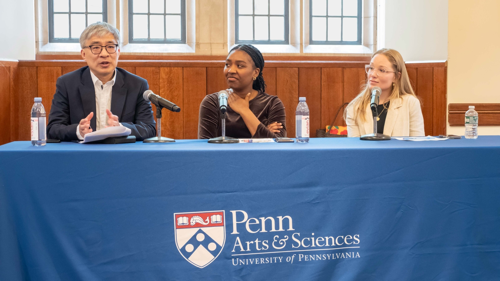 Guobin Yang, Adetobi Moses, and Liz Hallgren sitting at a table infront of windows. Yang speaks into a microphone, while Moses and Hallgren look towards him. Moses puts a hand on her chest. On the table are water bottles and a few microphones in stands positioned in front of the speakers. The tablecloth has the Penn logo, which includes the text: "Penn Arts & Sciences" and "University of Pennsylvania".