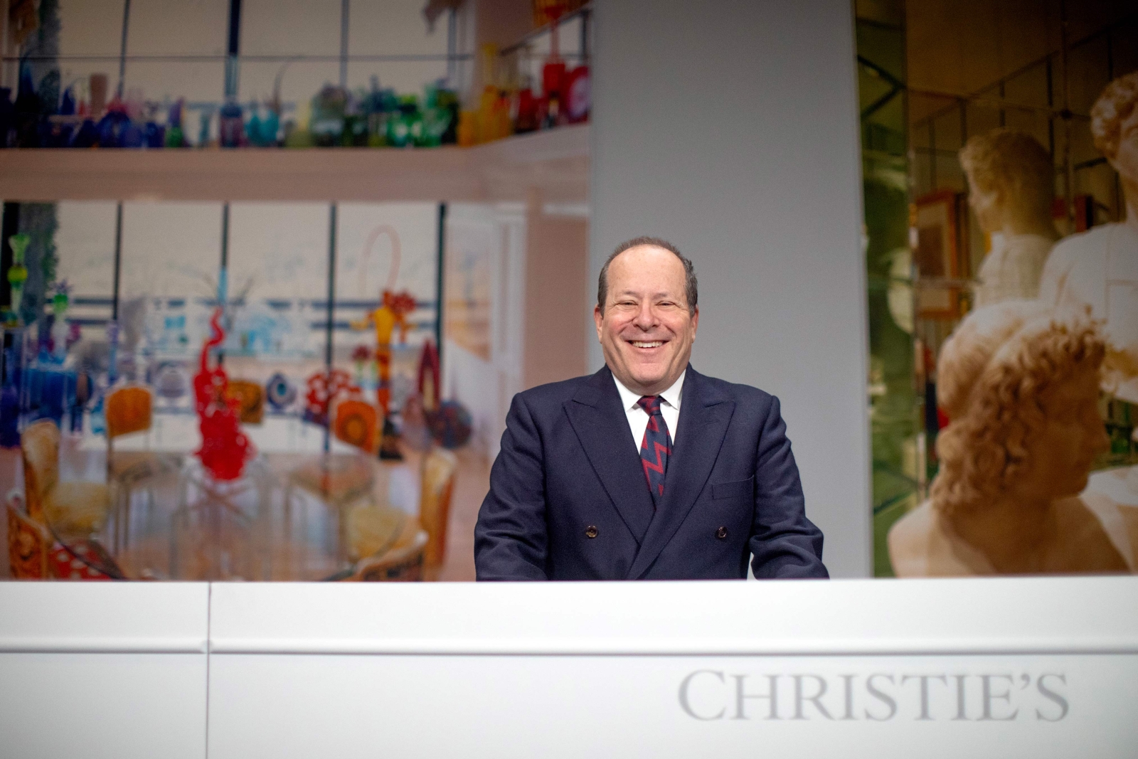 Marc Porter standing behind a large desk, smiling. The desk has the text "CHRISTIE'S" on the front, and in the background are large photos of sculptures in gallery spaces.
