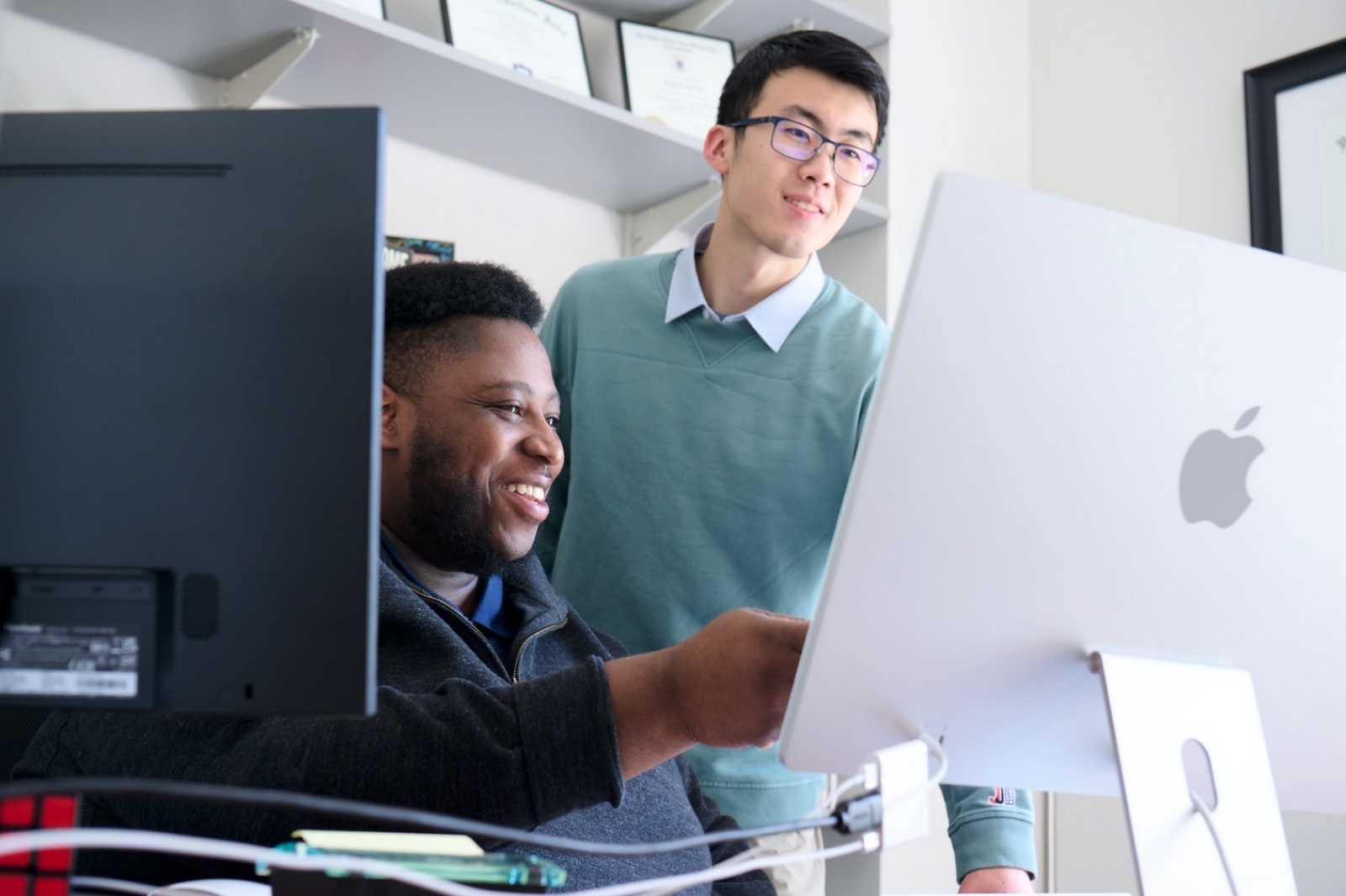 Two people looking at a computer screen, one sitting, one standing. The one sitting is pointing at the screen. 