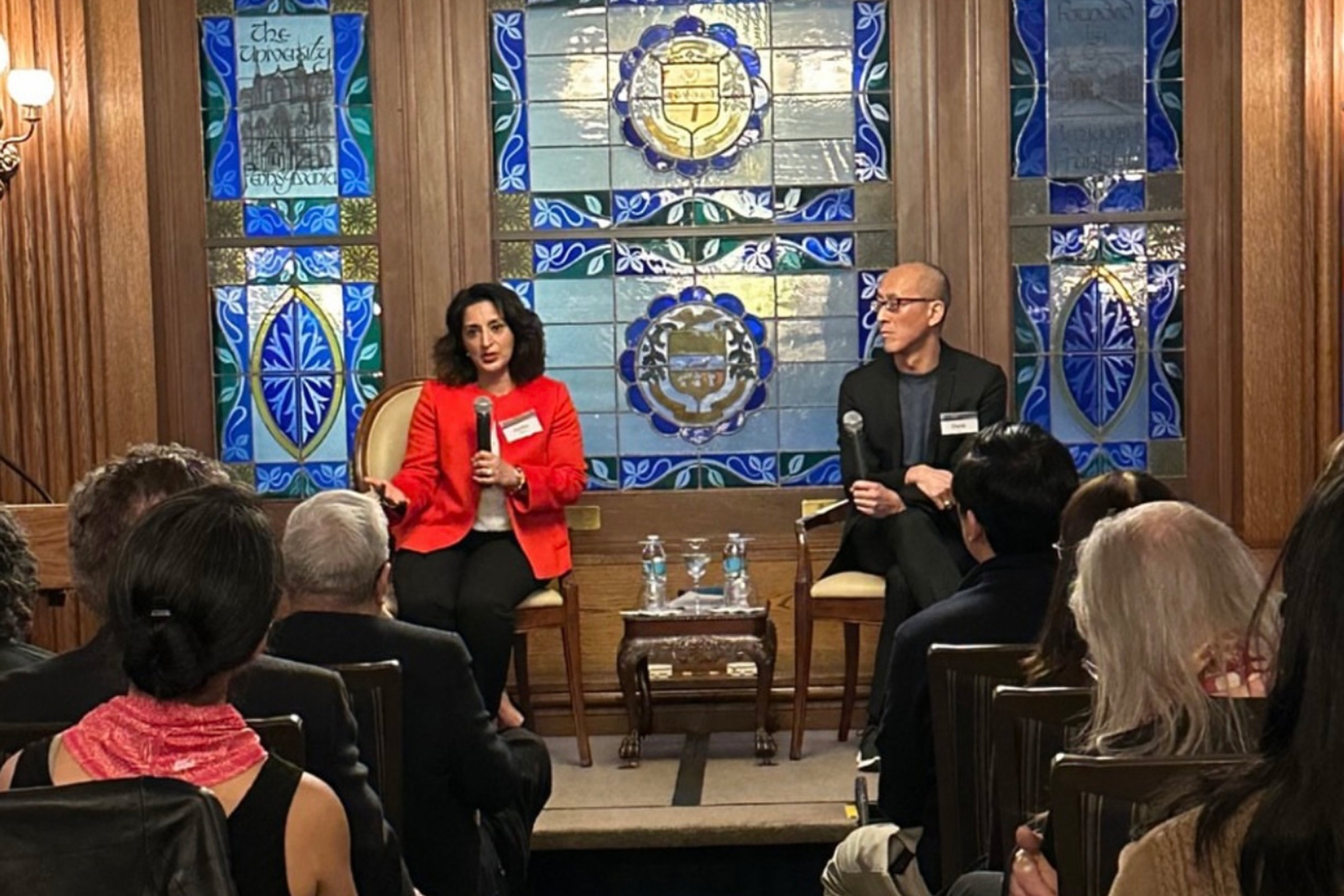 Fariha Khan and David Eng sitting on a stage addressing an audience. Khan speaks into a hand-held microphone while Eng listens intently. Behind them are three sets of stained glass panels.