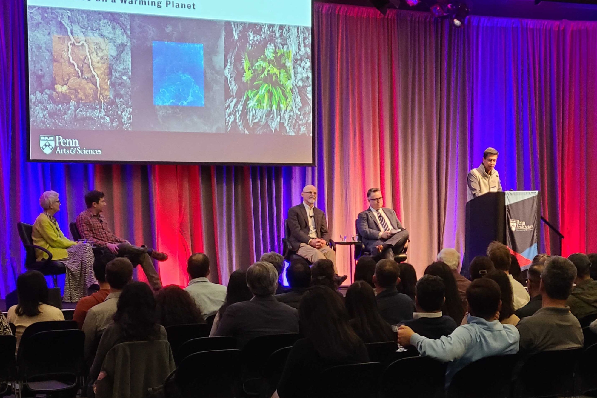 A crowd listening to several speakers on a stage. Four speakers are seated, while one person is at the podium. Behind them is a large screen with a slide projected reading: "BEN TALKS: PALO ALTO. Resilience on a Warming Planet." Also on the slide is the Penn arts and Sciences logo, as well as several images of nature scenes.