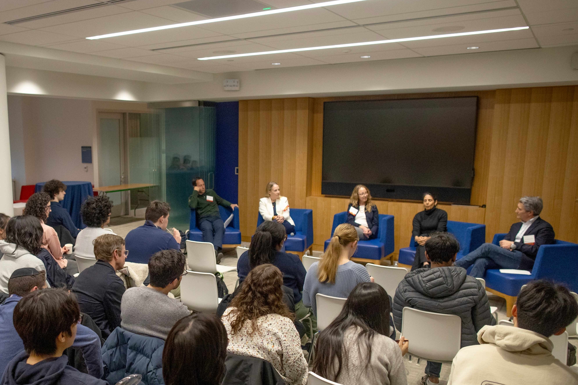 A group of speakers sitting in a arc in front of a room, speaking to an audience.