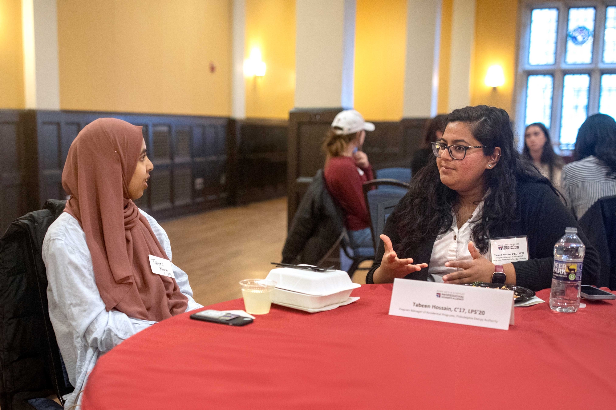 Tabeen Hossain speaking to a students at a table in a hall. Behind them is a nother group of students at a different table. Hossain speaks with her hands out, while the student listens intently. On the table is a cell phone, a cup of lemonade, a take out container, and a nametag with the text: "Tabeen Hossain C’17, LPS’20", as well as other type out of focus.