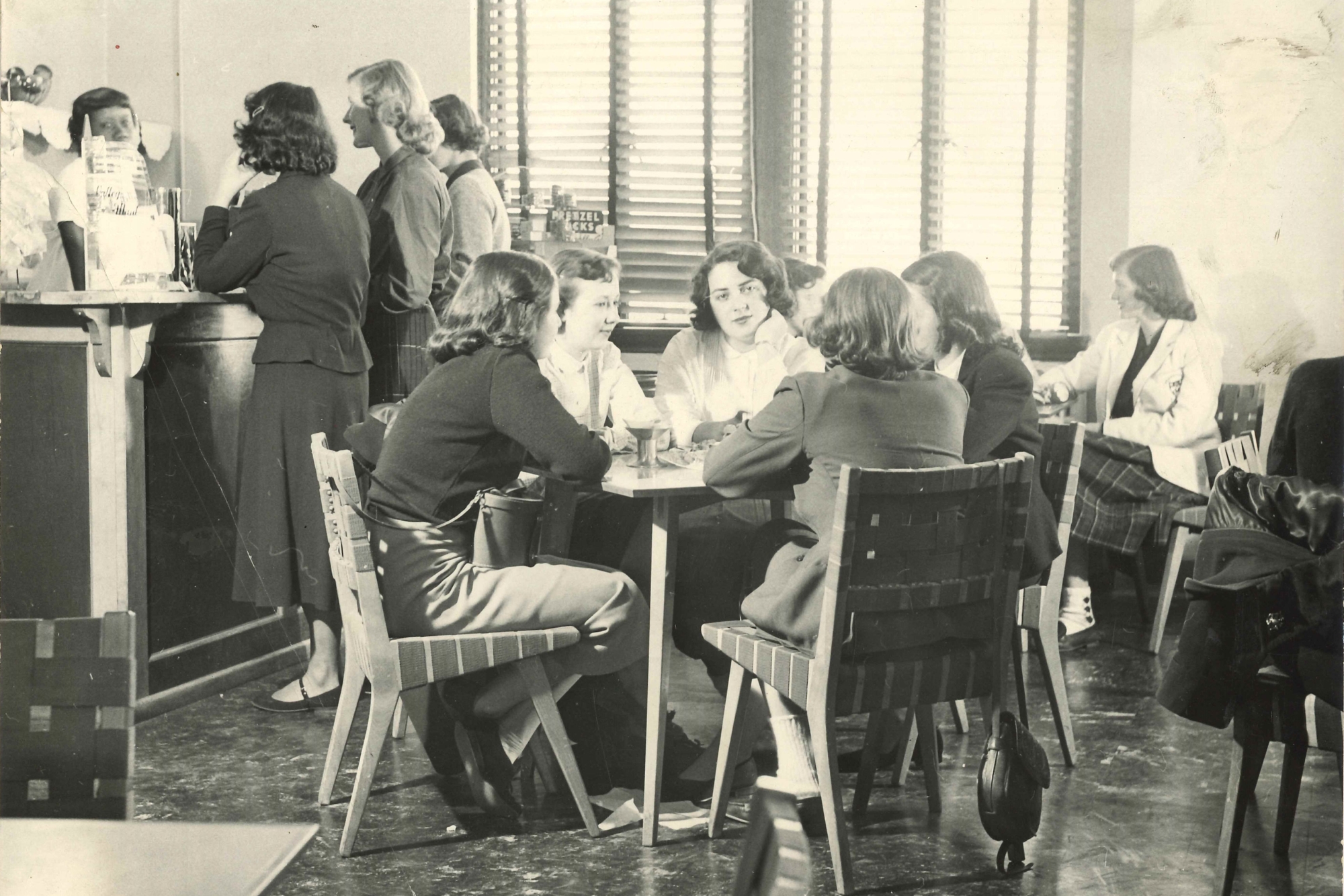 a group of women sitting at a table at a soda fountain. Behind them, a few others stand at a countertop, as well as a couple of people seated.