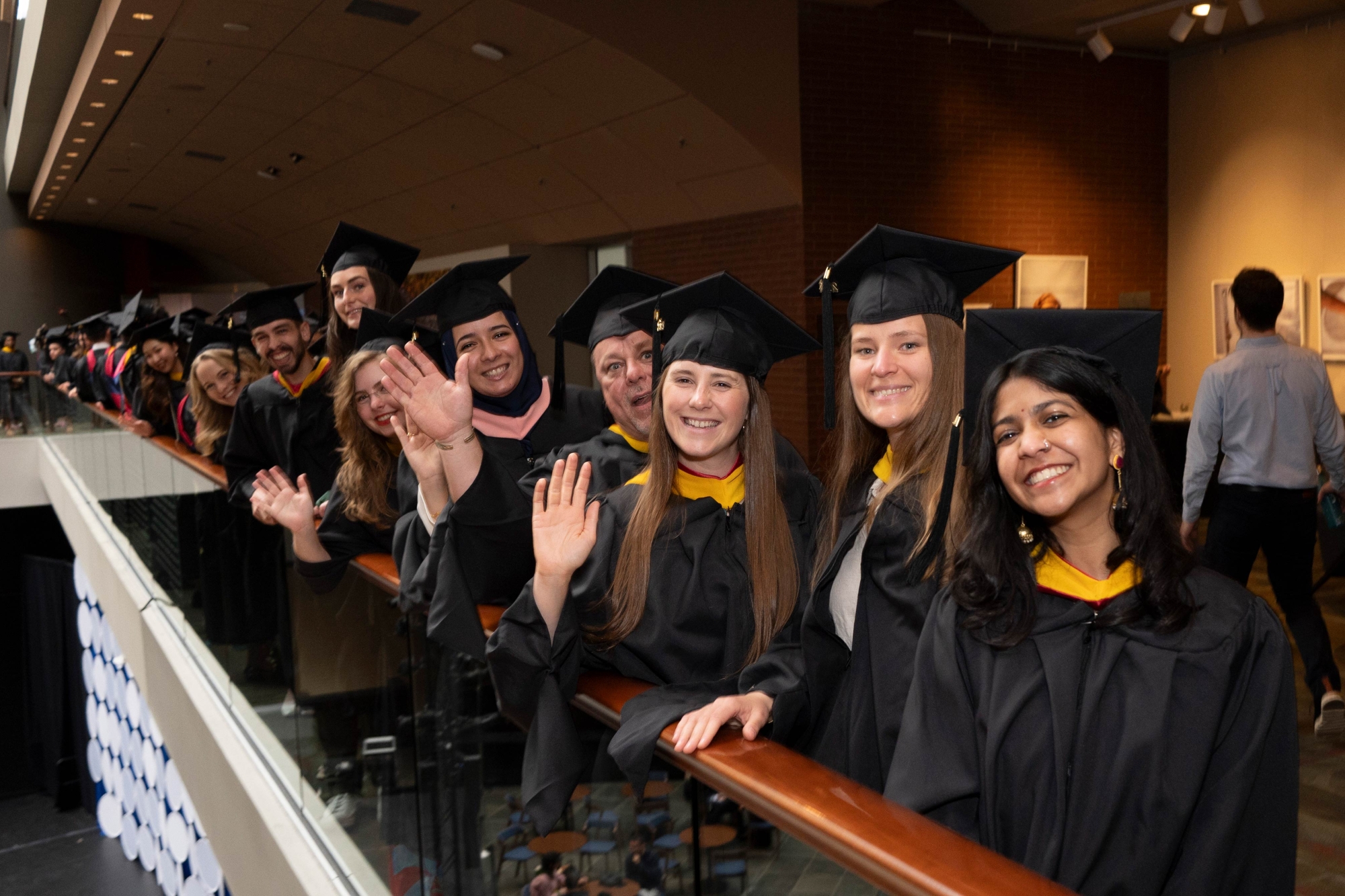 A group of excited college graduates in caps and gowns standing on a balcony, waving.