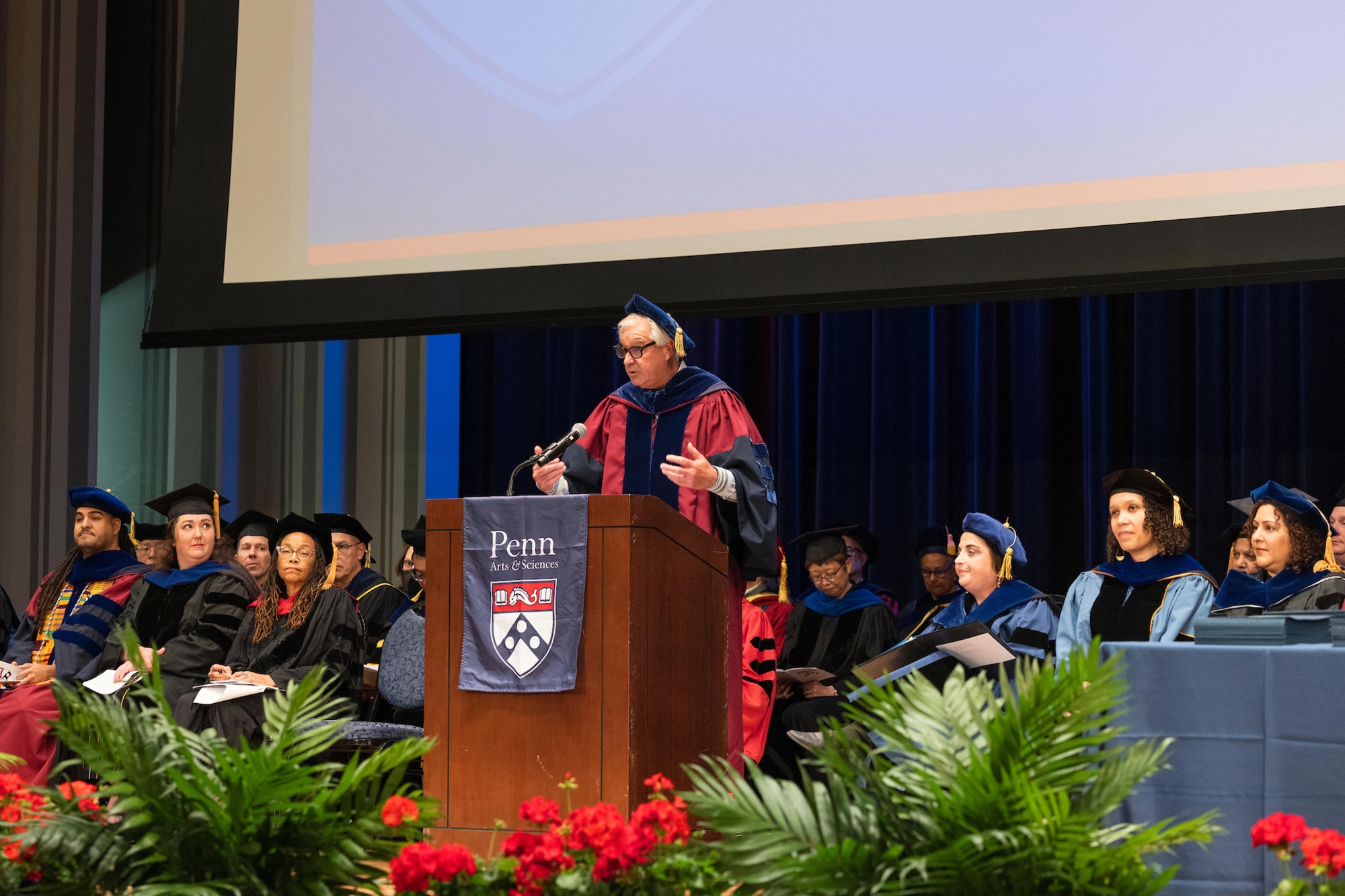 Dean Fluharty standing on a stage behind a in a graduation cap and gown, speaking. Behind him on stage are students, also in caps and gowns. In front of the stage are various plants, and a banner on the podium reads "Penn", and also features the Penn Shield.