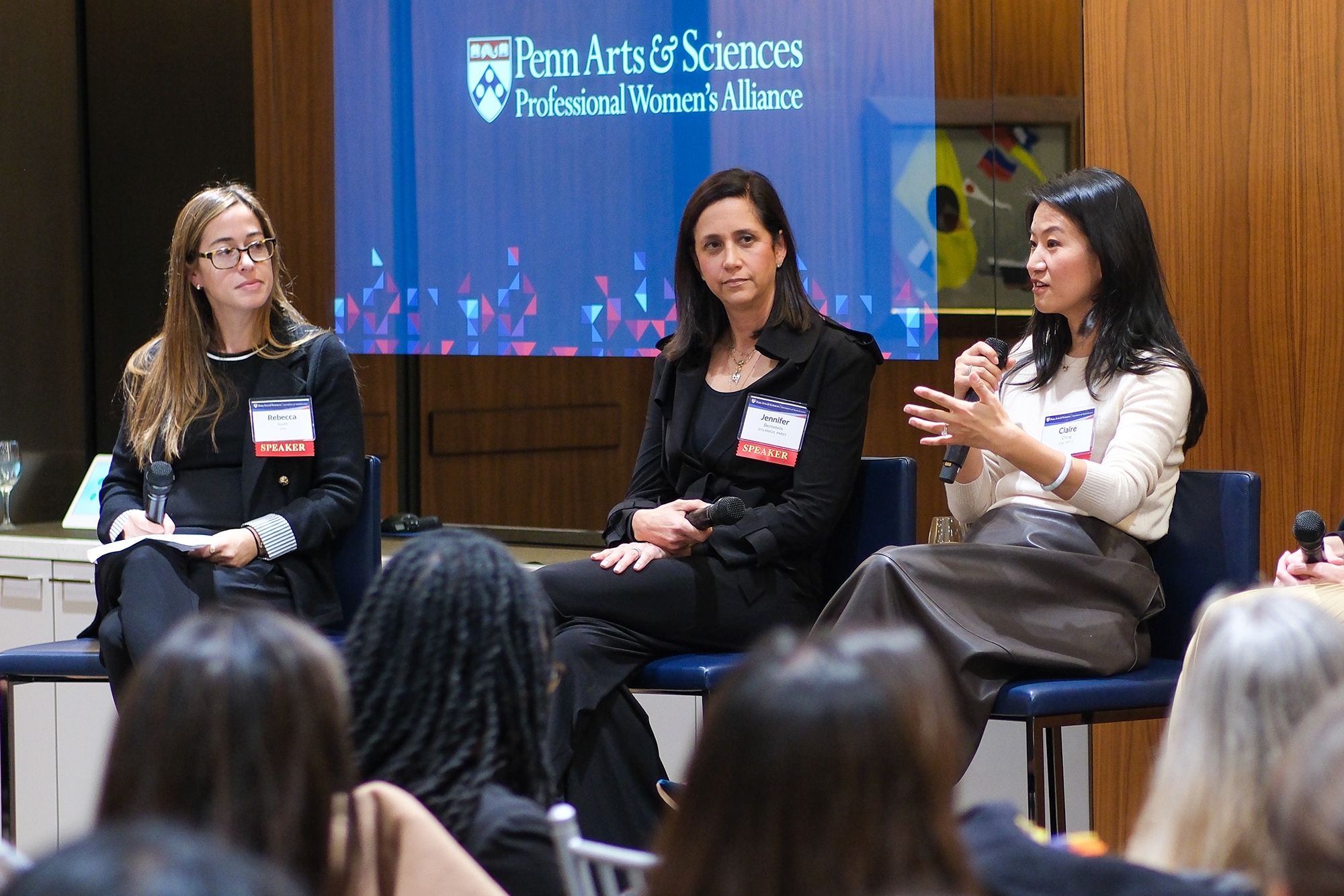 Three people sitting on a stage in tall chairs. One is holding a microphone, talking. Behind them a sign reads "Penn Arts & Sciences Professional Women's Alliance"