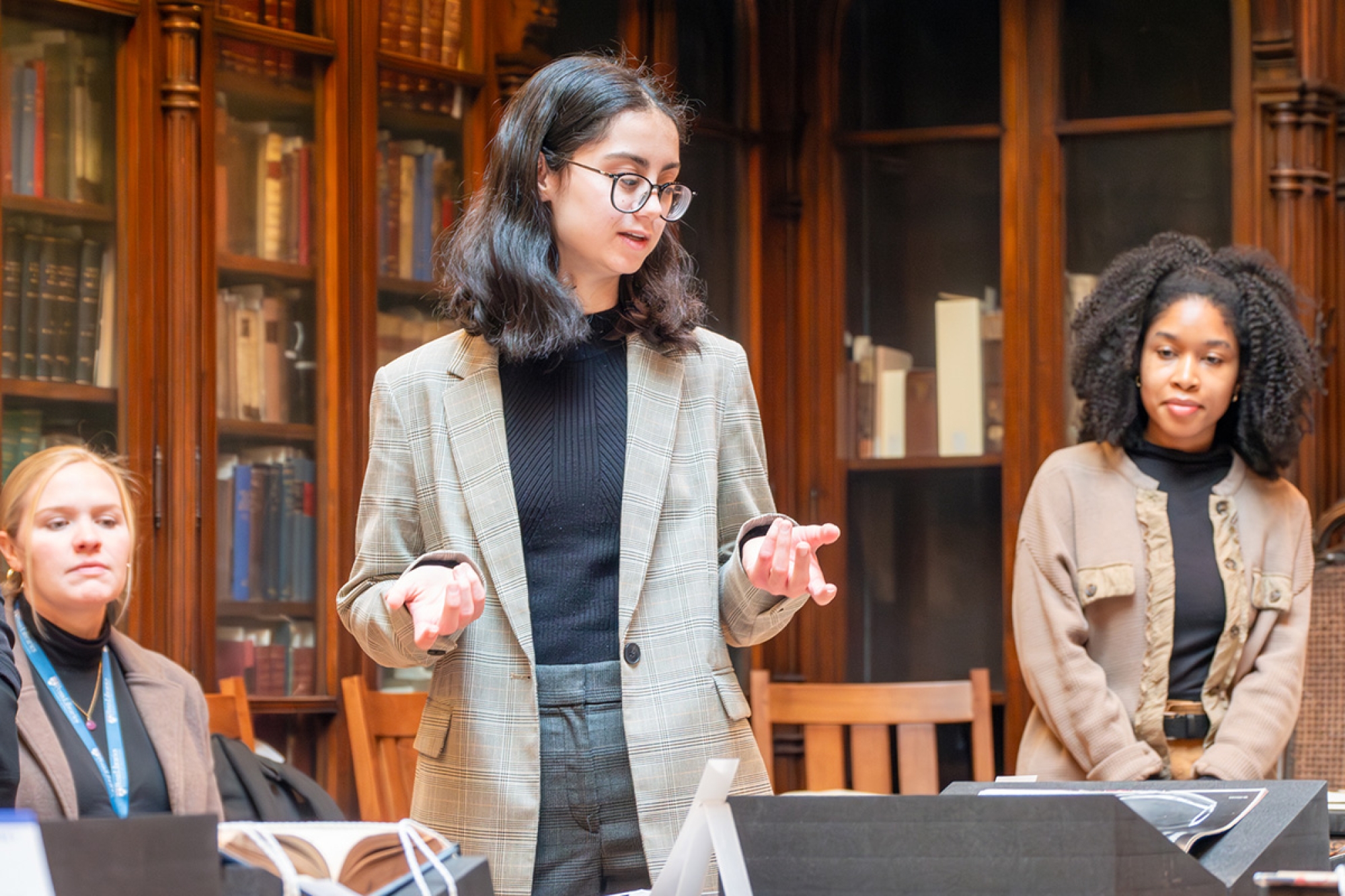 A person in a blazer and glasses standing up presenting materials, her hands outstretched wide. There is someone sitting to her left in the image, and someone else standing to her right. Behind them is wood paneling.