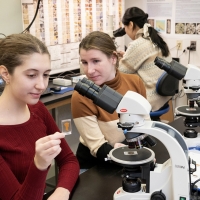 Two people sitting at a table with a microscope, with one person holding a slide and the other looking on. Other people and computers are in the background.