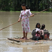 Three people on a raft. One, standing up, is slightly older, holding a stick to paddle across the water. The other two, younger children, site with their backpacks on their back. One is facing forward, the other facing back.