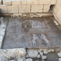 An excavation site. In the center are two mosaics on brown dirt, surrounded by a low rock wall and then higher limestone walls on three sides. 