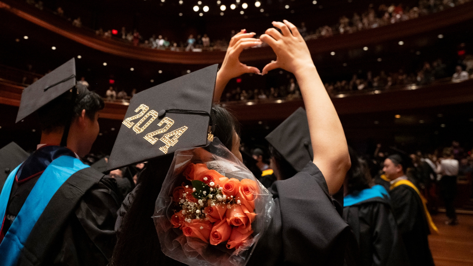 A person in a graduation cap with 2024 on the top making a heart shape with her hands. She has a bunch of roses draped on her shoulder.
