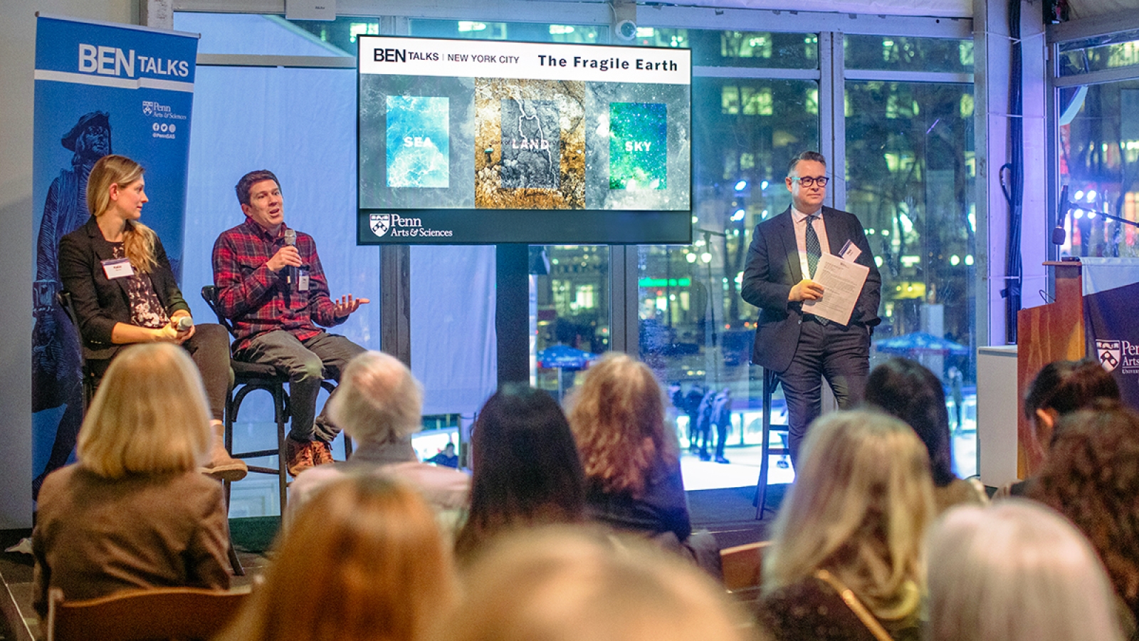 Katie Barott, Douglas Jerolmack, and Mark Trodden seated on a stage with a crowd in front of them. In the center of the stage is a television with the text "BEN TALKS NEW YORK CITY: The Fragile Earth" along with the words "Sea, Land, and Sky" on top of accompanying imagery. Jerolmack speaks into a hand-held microphone, and Trodden holds papers.