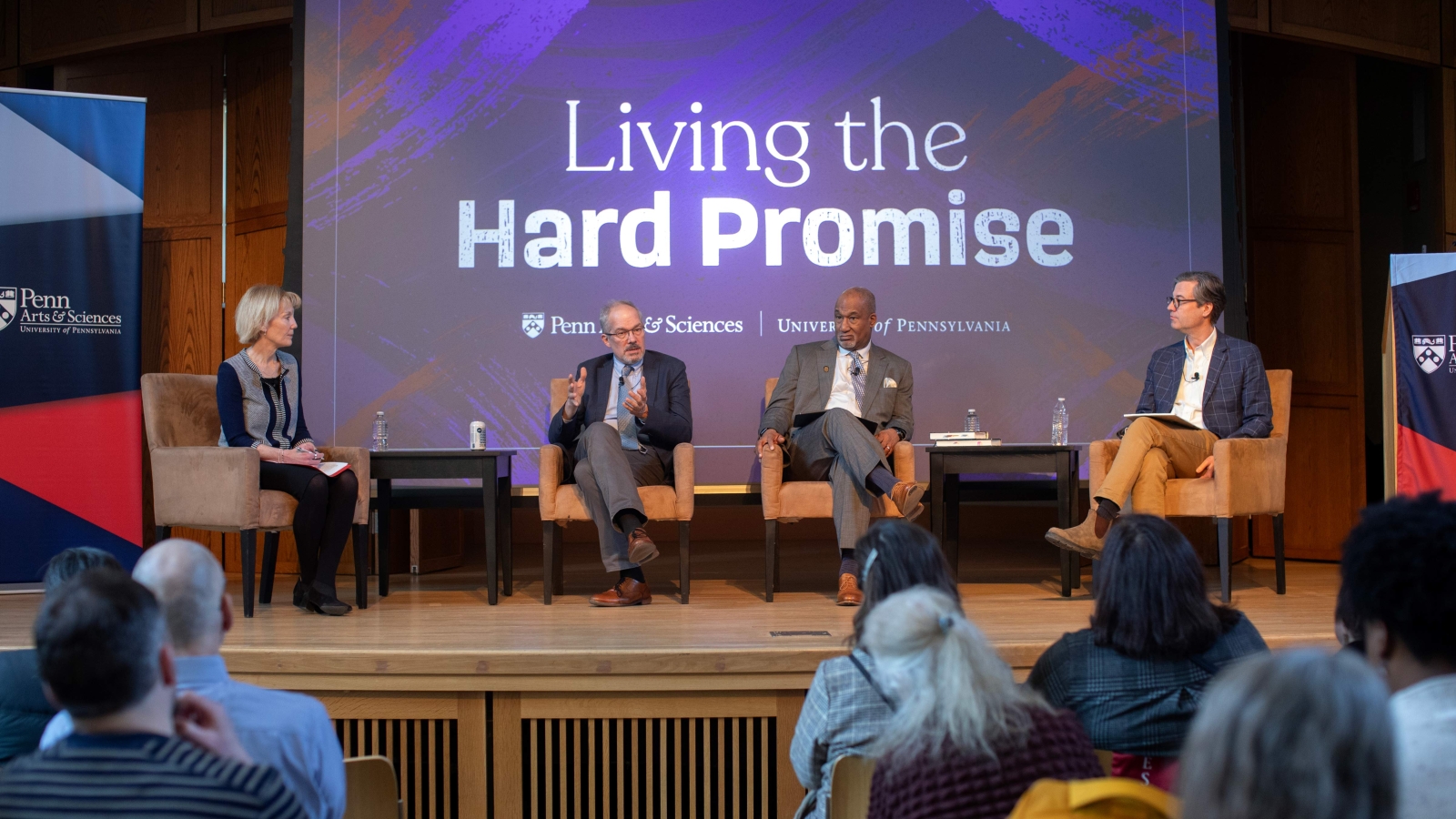 Laura Perna, Paul Sniegowski, Herman Bevears and Peter Struck seated on a stage in front of a crowd. Behind them is a screen displaying the words "Living the Hard Promise" as well as the Penn Arts and Sciences and University of Pennsylvania logos. Other panel members listen as Sniegowski speaks.