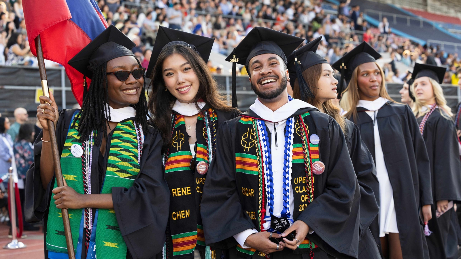 A group of students (Cystal Marshall, Rene Chen, Oscar Vasquez) standing in a crowded stadium. They are all wearing caps and gowns, and smiling. Marshall, on the left, holds a flag with the school colors.