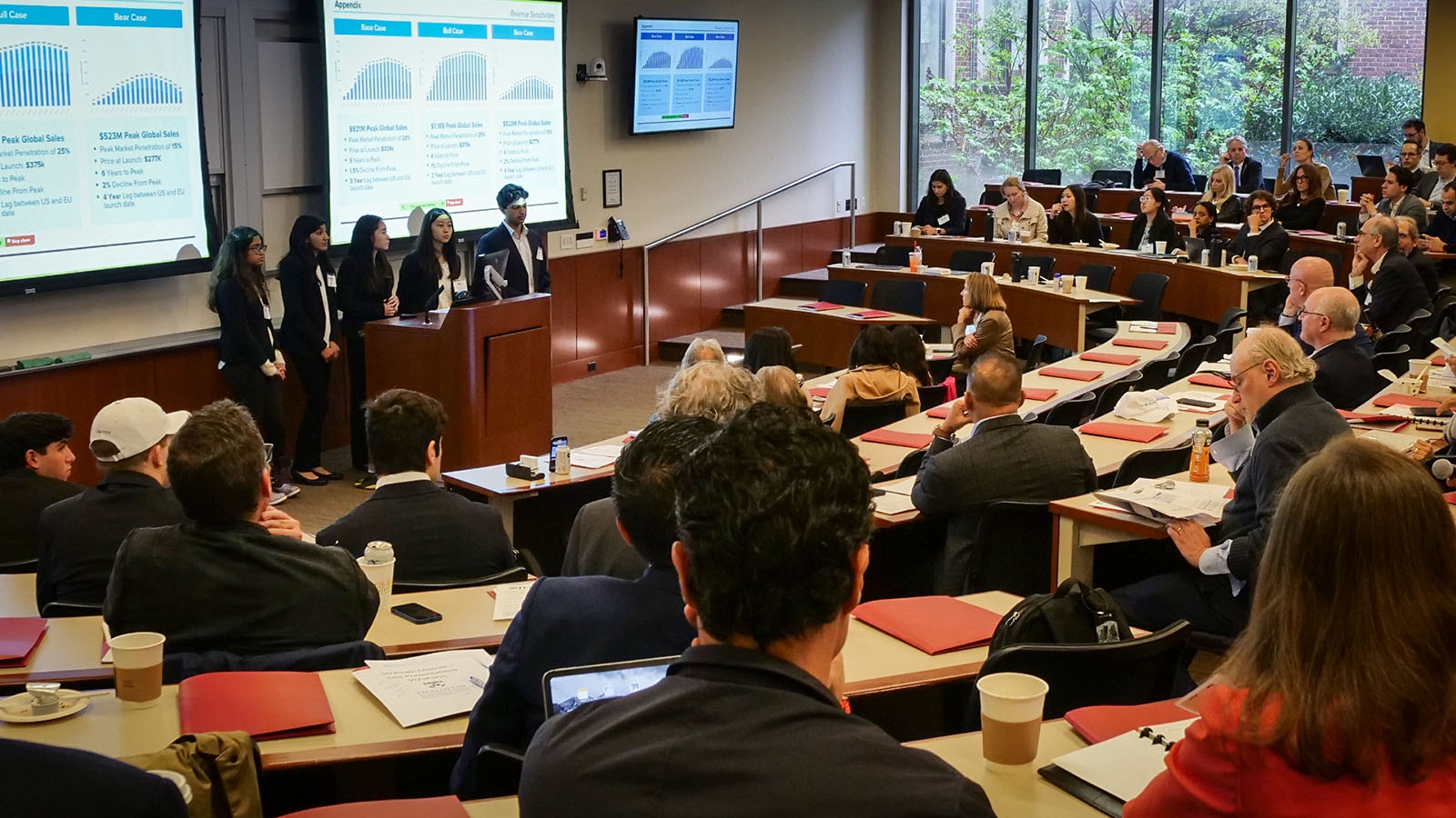 A group of students standing in the front of two large screens, presenting to a room of people sitting in an ampitheater-style classroom.