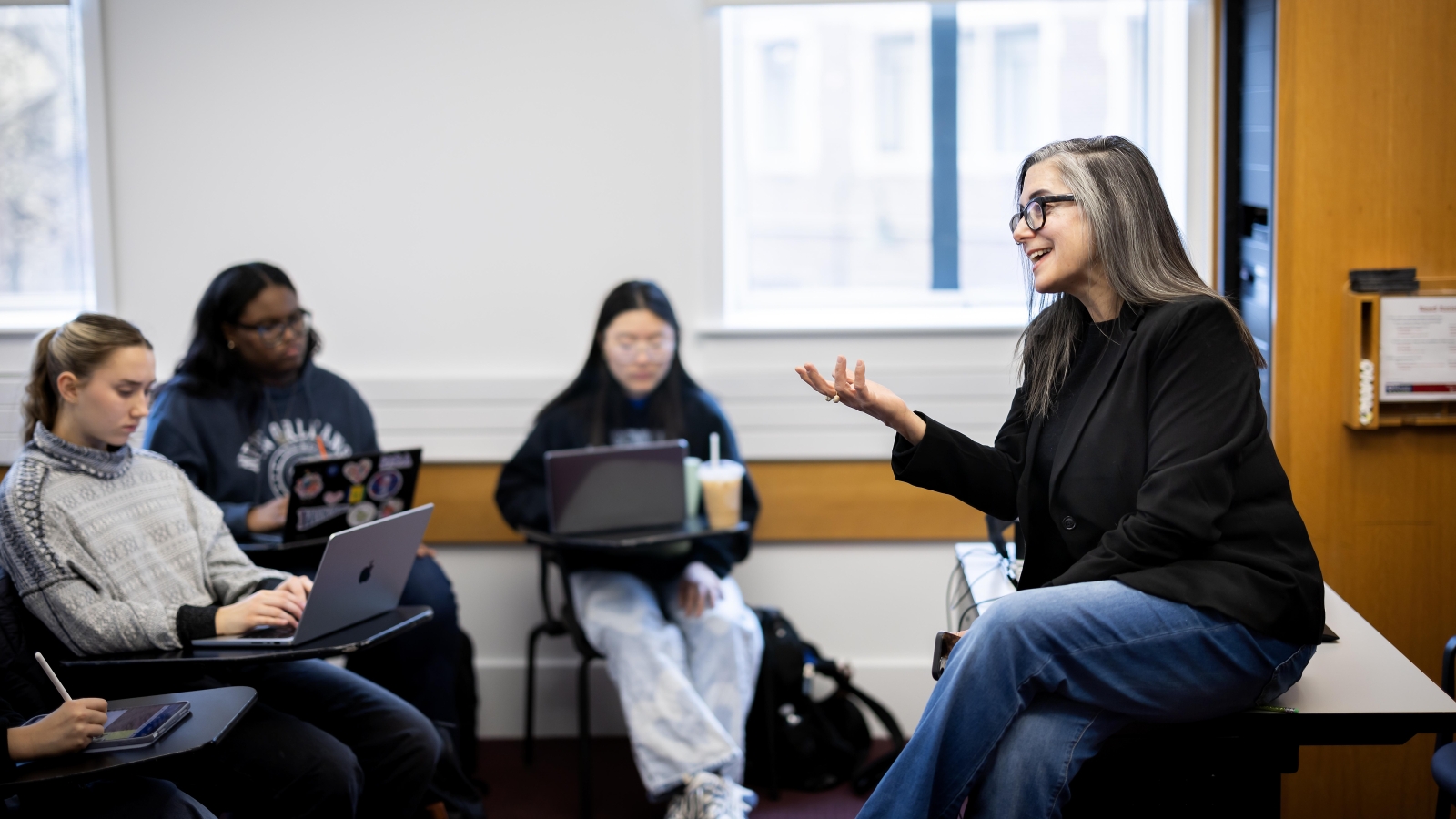 Melissa Jensen, who teaches in the English department, sitting on a desk talking to a room of students, three of whom are visible. 